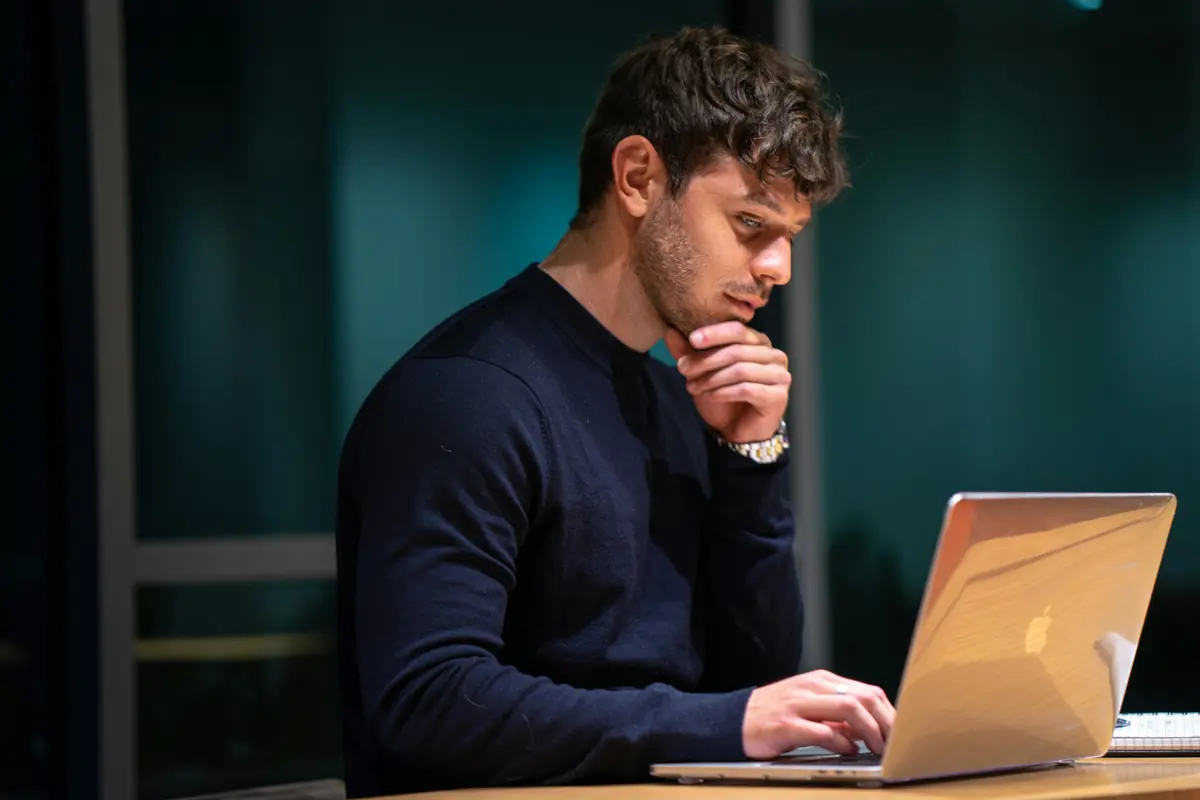 A man at a laptop with his chin in his hand, frowning at the screen