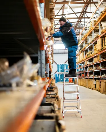 A warehouse worker on a stepladder reaching for a package on high racking