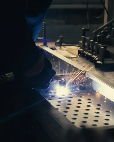 A welder working on a perforated steel sheet in a factory, sparks flying