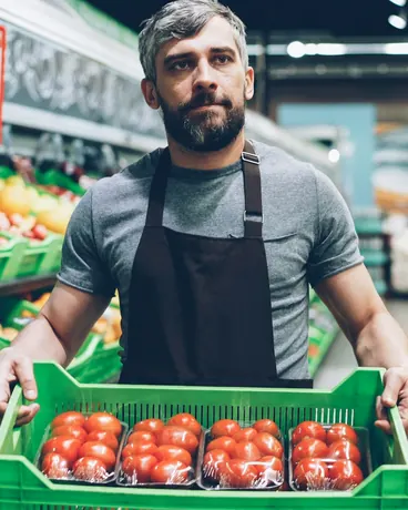 A grocery store worker in an apron carrying a crate of tomatoes through the produce aisle